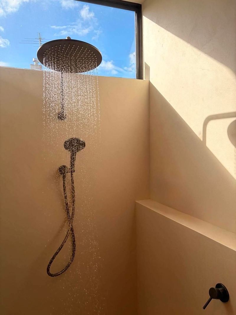 A seamless microcement shower in a warm sand tone with a rain shower head under a high window